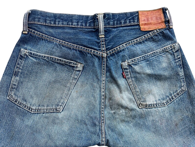 以下が含まれることがあります： 青いデニムジーンズの背面をクローズアップ。ジーンズには2つのバックポケット、右ポケットの小さな赤いタグ、茶色の革パッチが付いています。デニムは色あせ、使い古された外観をしています。