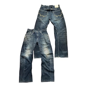 Può includere: Due paia di jeans in denim blu. Un paio è steso, l'altro è in piedi. Entrambi i paia hanno un lavaggio sbiadito, con usura visibile. I jeans presentano uno stile classico a cinque tasche, passanti per cintura e chiusura con bottone e cerniera.