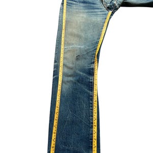 May include: Blue denim jeans with a faded wash and a distressed appearance. A yellow measuring tape runs along the length of the jeans. The jeans have a button fly and a patch on the knee.