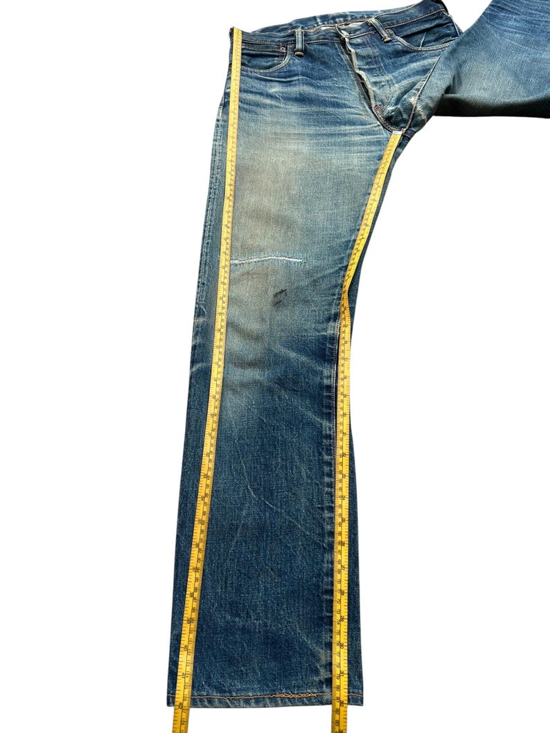 May include: Blue denim jeans with a faded wash and a distressed appearance. A yellow measuring tape runs along the length of the jeans. The jeans have a button fly and a patch on the knee.