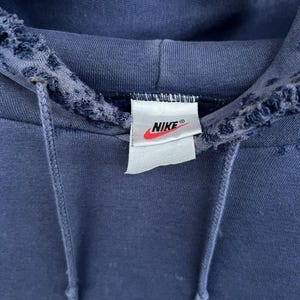Puede incluir: Una sudadera con capucha azul marino con una etiqueta blanca de Nike en la parte delantera. La sudadera tiene un aspecto desgastado con bordes deshilachados alrededor de la capucha.