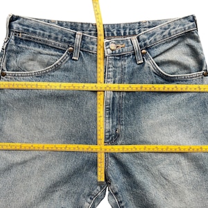 K&ouml;nnte beinhalten: Hellblaue Jeans mit klassischem F&uuml;nf-Taschen-Design. Ein gelbes Ma&szlig;band liegt &uuml;ber der Jeans, um die Bund- und Schrittl&auml;nge zu zeigen. Die Jeans hat einen Knopf- und Rei&szlig;verschluss.
