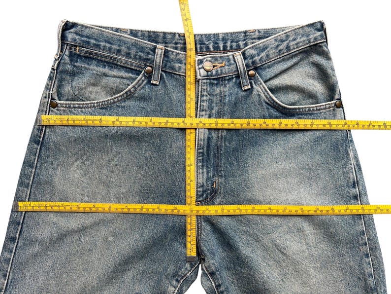 K&ouml;nnte beinhalten: Hellblaue Jeans mit klassischem F&uuml;nf-Taschen-Design. Ein gelbes Ma&szlig;band liegt &uuml;ber der Jeans, um die Bund- und Schrittl&auml;nge zu zeigen. Die Jeans hat einen Knopf- und Rei&szlig;verschluss.