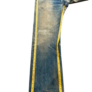 May include: Faded blue denim jeans with a distressed look, featuring a tear on the left leg. A yellow measuring tape is draped along the length of the jeans, providing a visual reference for size. The jeans have a button fly and classic five-pocket design.