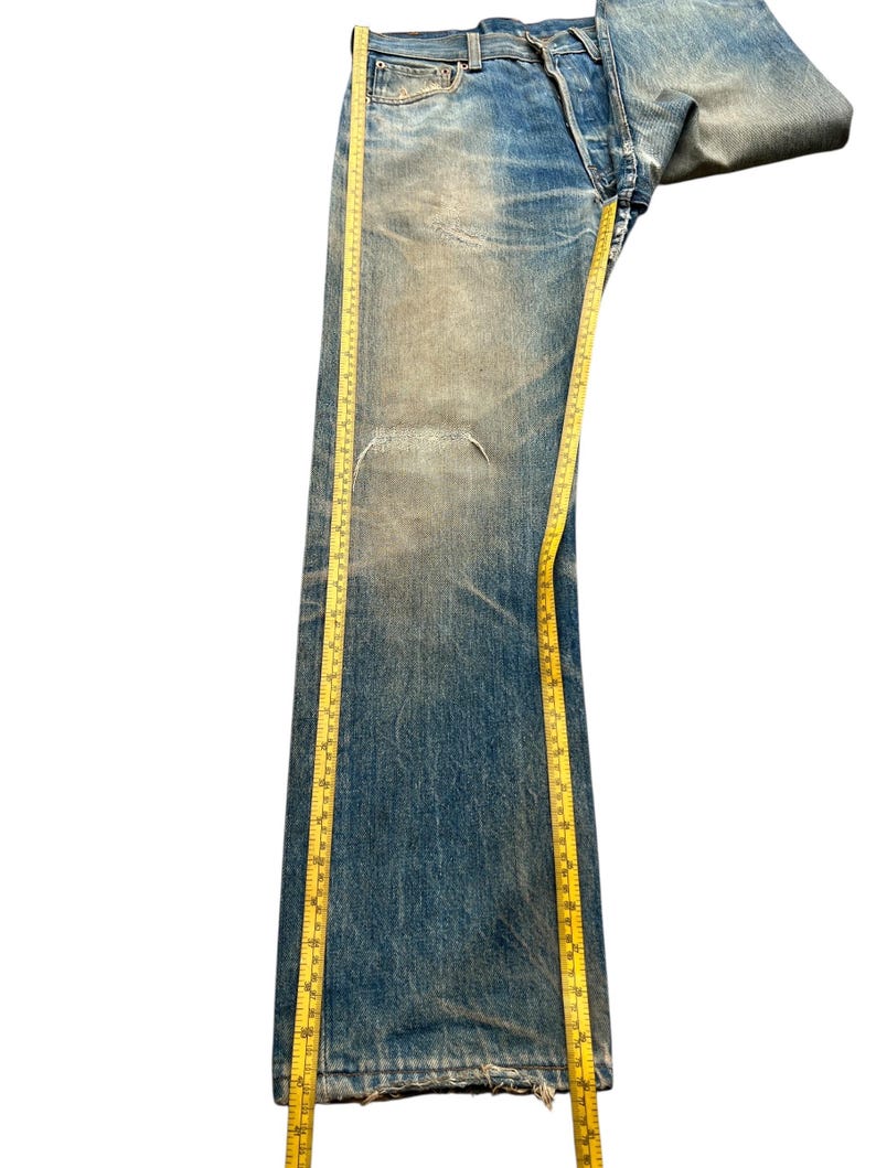 May include: Faded blue denim jeans with a distressed look, featuring a tear on the left leg. A yellow measuring tape is draped along the length of the jeans, providing a visual reference for size. The jeans have a button fly and classic five-pocket design.