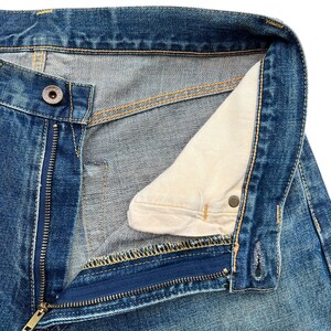 May include: Close-up of blue denim jeans with an open zipper and button closure. The jeans show visible stitching in yellow and a light-colored interior lining. The metal zipper and button are visible.