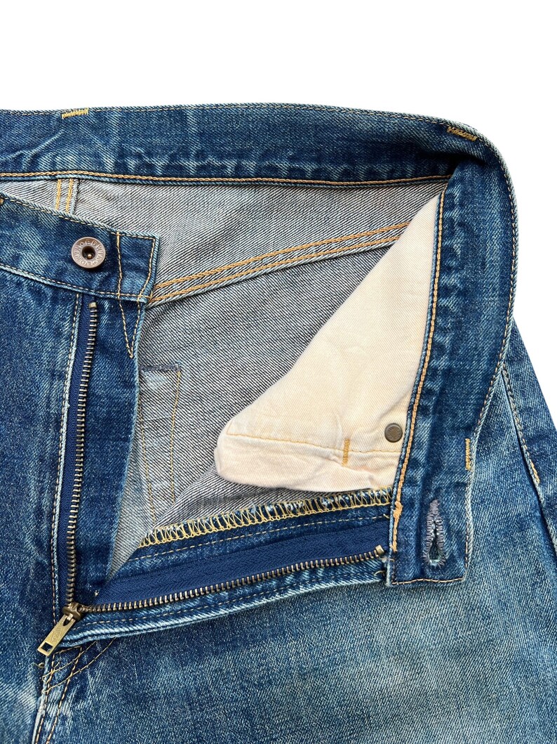 May include: Close-up of blue denim jeans with an open zipper and button closure. The jeans show visible stitching in yellow and a light-colored interior lining. The metal zipper and button are visible.