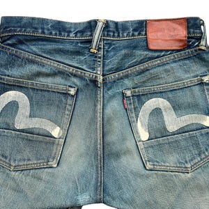 May include: Close-up of the back of blue denim jeans. The jeans have white painted designs on the back pockets and a brown leather patch. The denim is faded and distressed, with a classic five-pocket design.