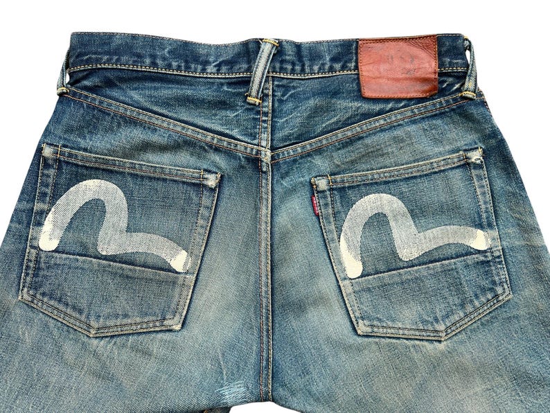 May include: Close-up of the back of blue denim jeans. The jeans have white painted designs on the back pockets and a brown leather patch. The denim is faded and distressed, with a classic five-pocket design.