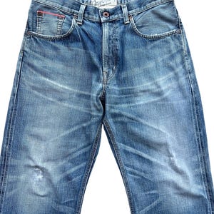 Pu&ograve; includere: Jeans di denim blu sbiadito con un classico design a cinque tasche. Presenta una chiusura con bottone e un piccolo accento rosso sulla tasca anteriore. I jeans hanno un aspetto invecchiato con sbiadimento.
