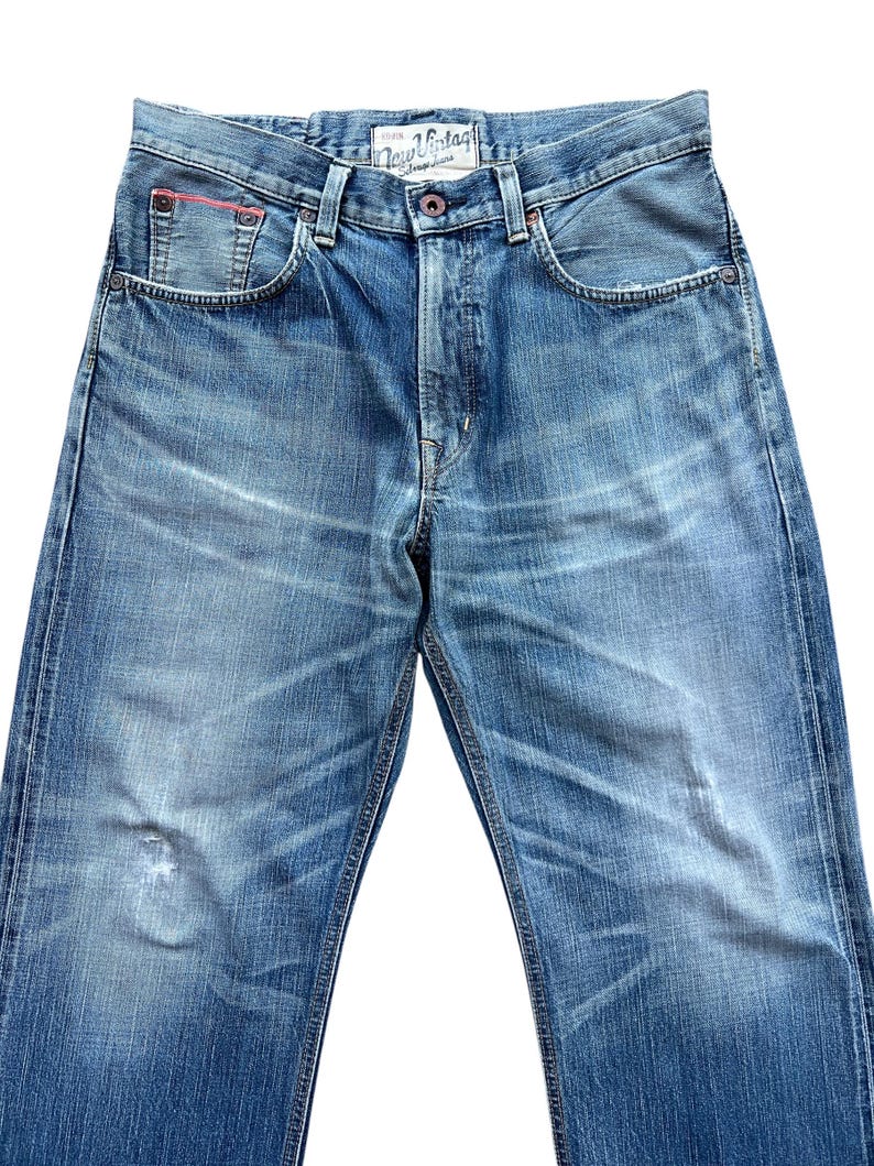 Pu&ograve; includere: Jeans di denim blu sbiadito con un classico design a cinque tasche. Presenta una chiusura con bottone e un piccolo accento rosso sulla tasca anteriore. I jeans hanno un aspetto invecchiato con sbiadimento.