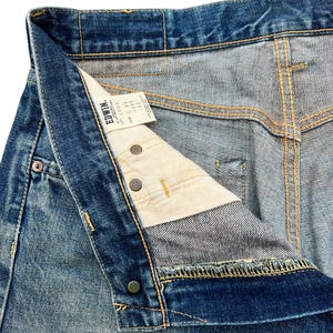 May include: Close-up of blue denim jeans with a button fly. The jeans have a faded wash and visible stitching in yellow thread. A white tag with black text is visible inside the waistband. The jeans are unbuttoned, showing the interior construction.
