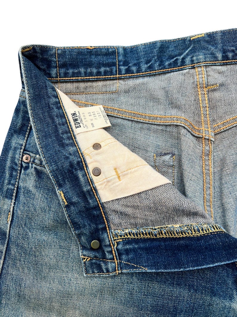 May include: Close-up of blue denim jeans with a button fly. The jeans have a faded wash and visible stitching in yellow thread. A white tag with black text is visible inside the waistband. The jeans are unbuttoned, showing the interior construction.