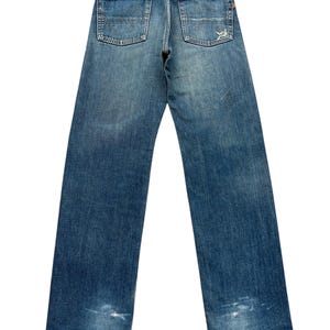 Puede incluir: Vaqueros de mezclilla azul descolorido con un dise&ntilde;o cl&aacute;sico de cinco bolsillos. Los vaqueros muestran signos de desgaste, incluyendo &aacute;reas desgastadas y un parche de cuero. Los bolsillos traseros tienen un dise&ntilde;o cosido. Los vaqueros son de estilo vintage.