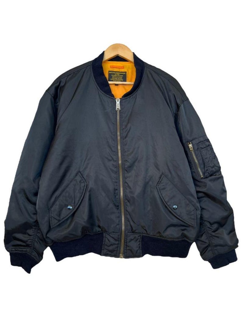 May include: A black bomber jacket with a zip-up front, two front pockets, and a pocket on the left sleeve. The jacket has a dark blue collar and cuffs, and a bright orange lining. The jacket is hanging on a wooden hanger.