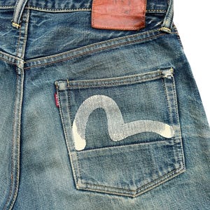 May include: Close-up of blue denim jeans with a white painted design on the back pocket. The jeans have a brown leather patch and a red tag. The denim has a worn, faded appearance.