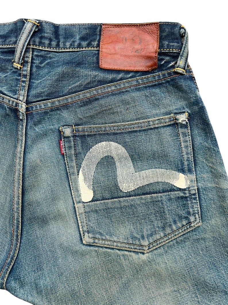 May include: Close-up of blue denim jeans with a white painted design on the back pocket. The jeans have a brown leather patch and a red tag. The denim has a worn, faded appearance.