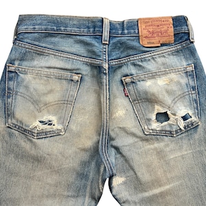May include: Close-up of vintage blue denim jeans with distressed details. The jeans feature worn patches, a faded wash, and a leather Levi Strauss & Co. label. The back pockets show the iconic Levi's stitching and a small red tag.