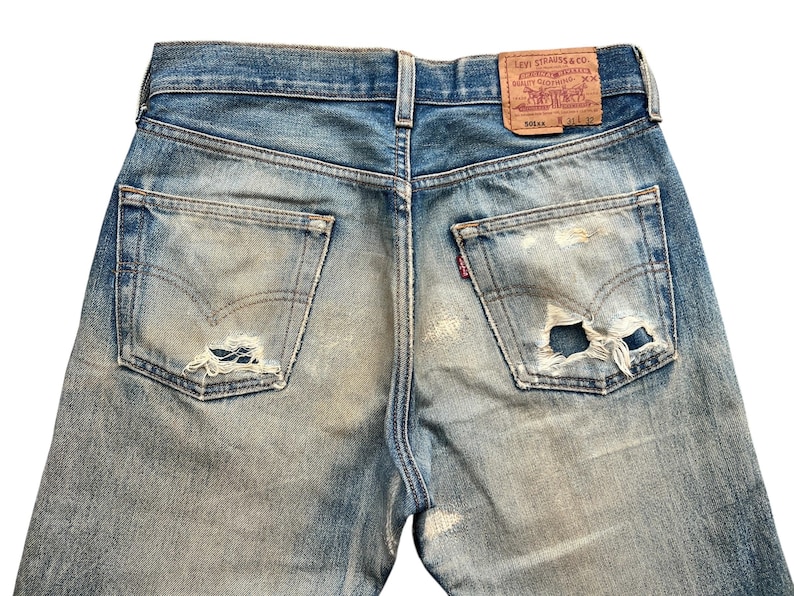 May include: Close-up of vintage blue denim jeans with distressed details. The jeans feature worn patches, a faded wash, and a leather Levi Strauss & Co. label. The back pockets show the iconic Levi's stitching and a small red tag.