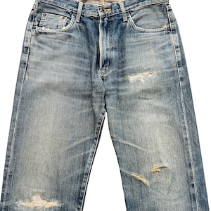 May include: Faded blue denim jeans with distressed details, including rips and worn patches. The jeans have a classic five-pocket design, a button closure, and a zip fly. The denim has a vintage wash.
