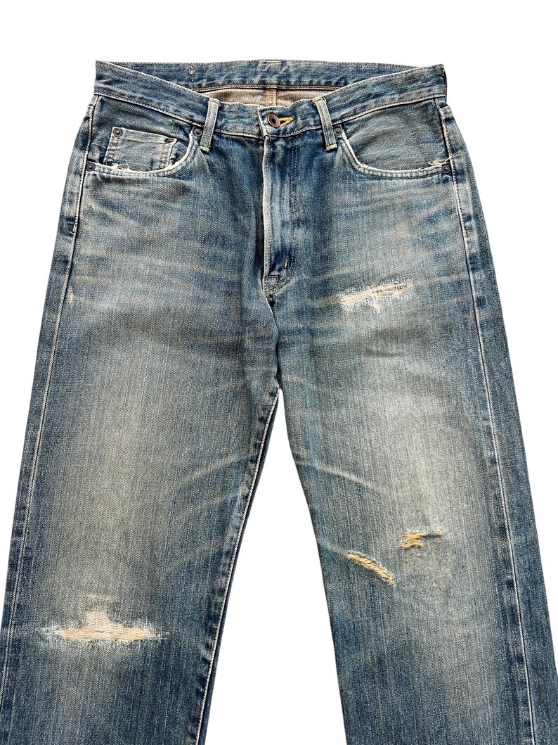 May include: Faded blue denim jeans with distressed details, including rips and worn patches. The jeans have a classic five-pocket design, a button closure, and a zip fly. The denim has a vintage wash.