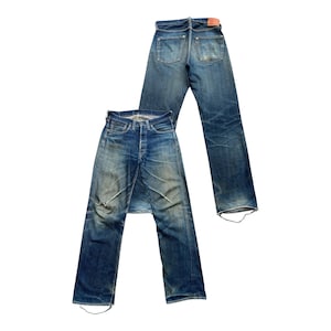 Może przedstawiać: Dwie pary dżins&oacute;w z niebieskiego denimu w stylu vintage. Jedna para leży płasko, pokazując prz&oacute;d, z zapięciem na guziki i przetarciami. Druga para jest za nią, pokazując tylne kieszenie i czerwoną sk&oacute;rzaną naszywkę.