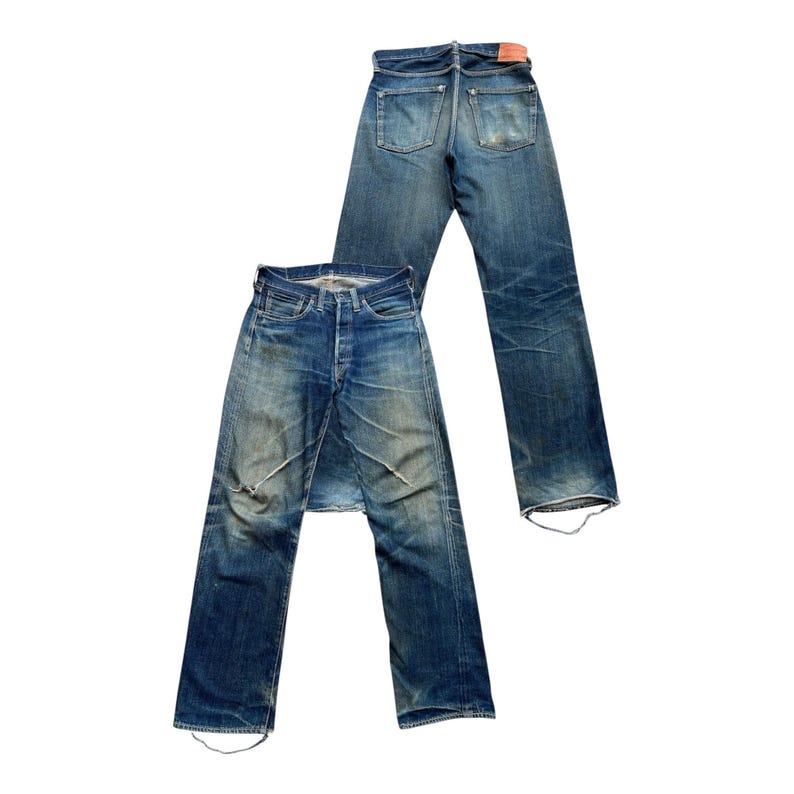 Może przedstawiać: Dwie pary dżins&oacute;w z niebieskiego denimu w stylu vintage. Jedna para leży płasko, pokazując prz&oacute;d, z zapięciem na guziki i przetarciami. Druga para jest za nią, pokazując tylne kieszenie i czerwoną sk&oacute;rzaną naszywkę.
