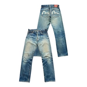 May include: Two pairs of blue denim jeans. One pair is laid flat, showing the front with button fly and distressed areas. The other pair is upright, displaying the back pockets with a white seagull design and a red leather patch.