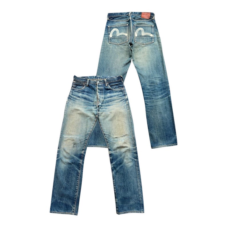 May include: Two pairs of blue denim jeans. One pair is laid flat, showing the front with button fly and distressed areas. The other pair is upright, displaying the back pockets with a white seagull design and a red leather patch.