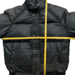 May include: Black leather puffer jacket with a high collar, button closure, and two front pockets. The jacket has a quilted design and a ribbed hem. A yellow measuring tape is laid across the jacket to measure its dimensions.