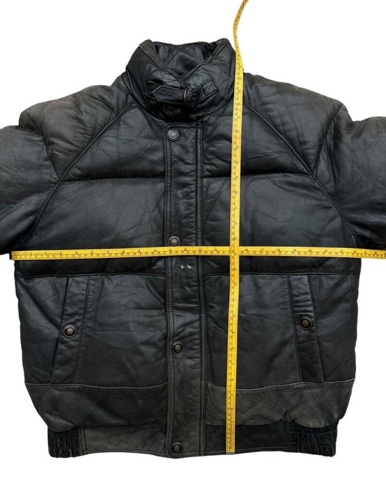 May include: Black leather puffer jacket with a high collar, button closure, and two front pockets. The jacket has a quilted design and a ribbed hem. A yellow measuring tape is laid across the jacket to measure its dimensions.