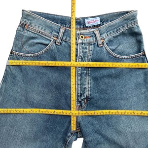 May include: Blue denim Wrangler jeans with a classic five-pocket design. The jeans are shown with a yellow measuring tape, indicating potential measurements. The Wrangler label is visible on the back pocket.