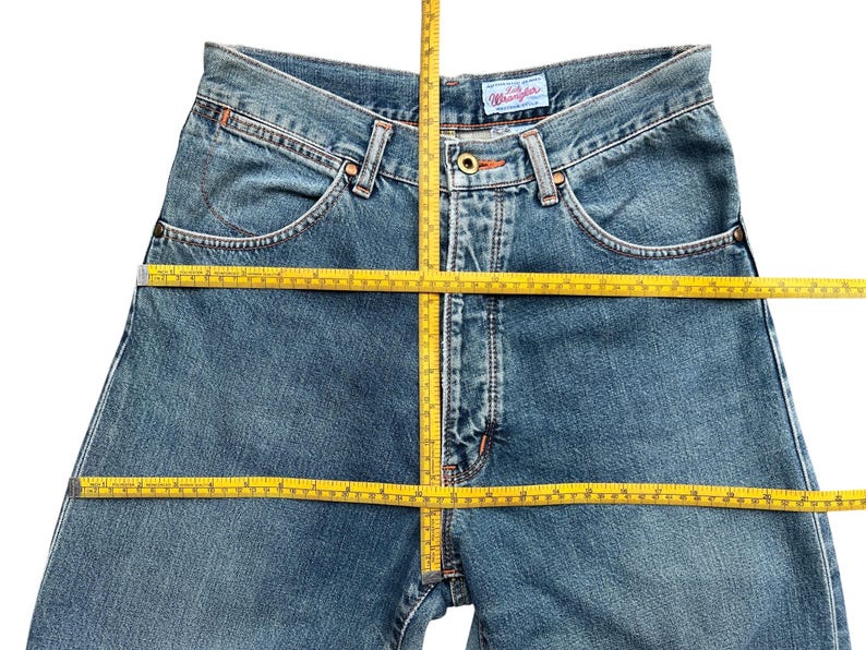May include: Blue denim Wrangler jeans with a classic five-pocket design. The jeans are shown with a yellow measuring tape, indicating potential measurements. The Wrangler label is visible on the back pocket.