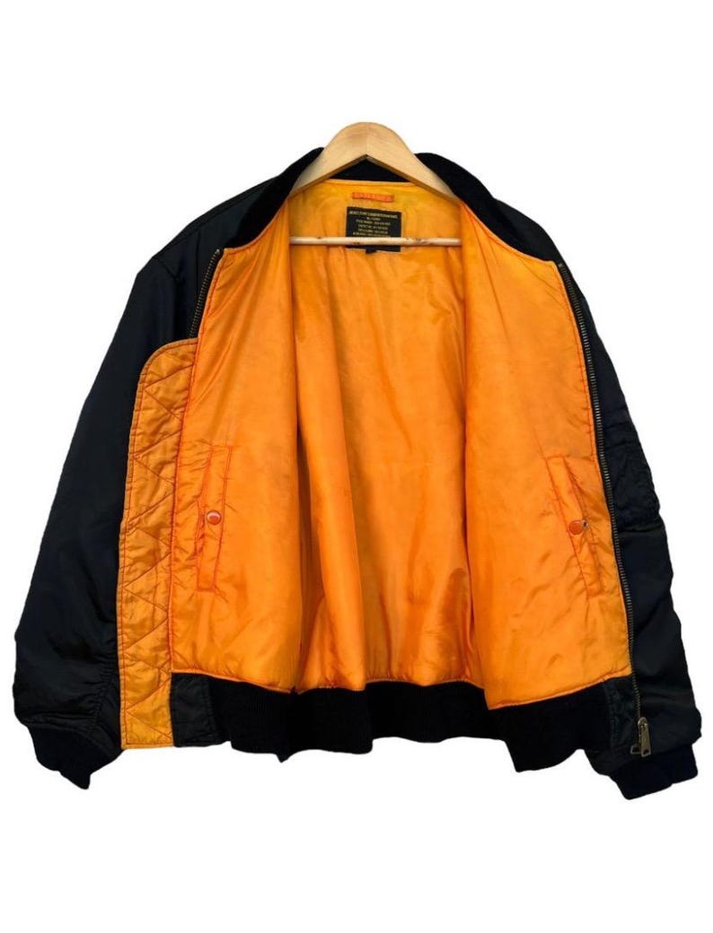 May include: Black bomber jacket with an orange interior. The jacket is hanging on a wooden hanger. The interior lining is a bright orange color, and the exterior is black. The jacket has a zipper closure and pockets.