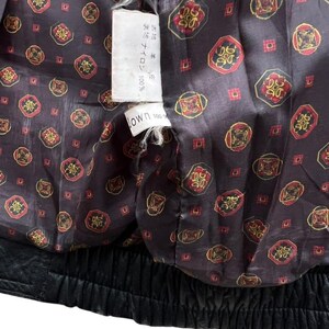May include: Close-up of the inside of a black leather jacket. The lining is a dark brown fabric with a repeating pattern of red and gold circular designs. A white tag with Japanese text is visible.