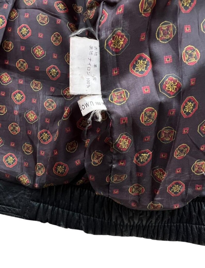 May include: Close-up of the inside of a black leather jacket. The lining is a dark brown fabric with a repeating pattern of red and gold circular designs. A white tag with Japanese text is visible.