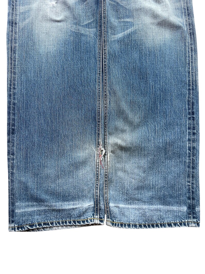 Pu&ograve; includere: Primo piano di un paio di jeans blu in denim. I jeans mostrano segni di usura e sbiadimento, con un'area usurata vicino al fondo. Le cuciture sono visibili lungo le cuciture e l'orlo. L'aspetto generale &egrave; casual e usurato.