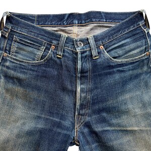Może przedstawiać: Zbliżenie niebieskich dżins&oacute;w. Dżinsy mają rozporek na guziki, klasyczny pięciokieszeniowy design i miedziane nity. Denim wykazuje blaknięcie i zużycie, z jasnoniebieskim praniem. Dżinsy to klasyczny styl.