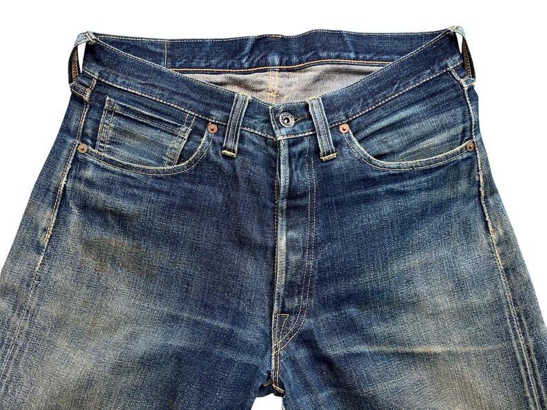 Może przedstawiać: Zbliżenie niebieskich dżins&oacute;w. Dżinsy mają rozporek na guziki, klasyczny pięciokieszeniowy design i miedziane nity. Denim wykazuje blaknięcie i zużycie, z jasnoniebieskim praniem. Dżinsy to klasyczny styl.
