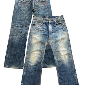 Puede incluir: Dos pares de vaqueros de mezclilla azul. Uno muestra los bolsillos traseros con detalles de costura rojos. El otro, al frente, presenta un aspecto desgastado con un lavado descolorido y una rodilla rasgada. Los vaqueros tienen un diseño clásico de cinco bolsillos.