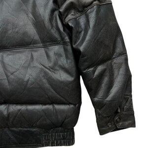 May include: Close-up of a black leather puffer jacket. The jacket features a quilted design, a ribbed hem, and a buttoned cuff. The leather has a slightly distressed appearance.