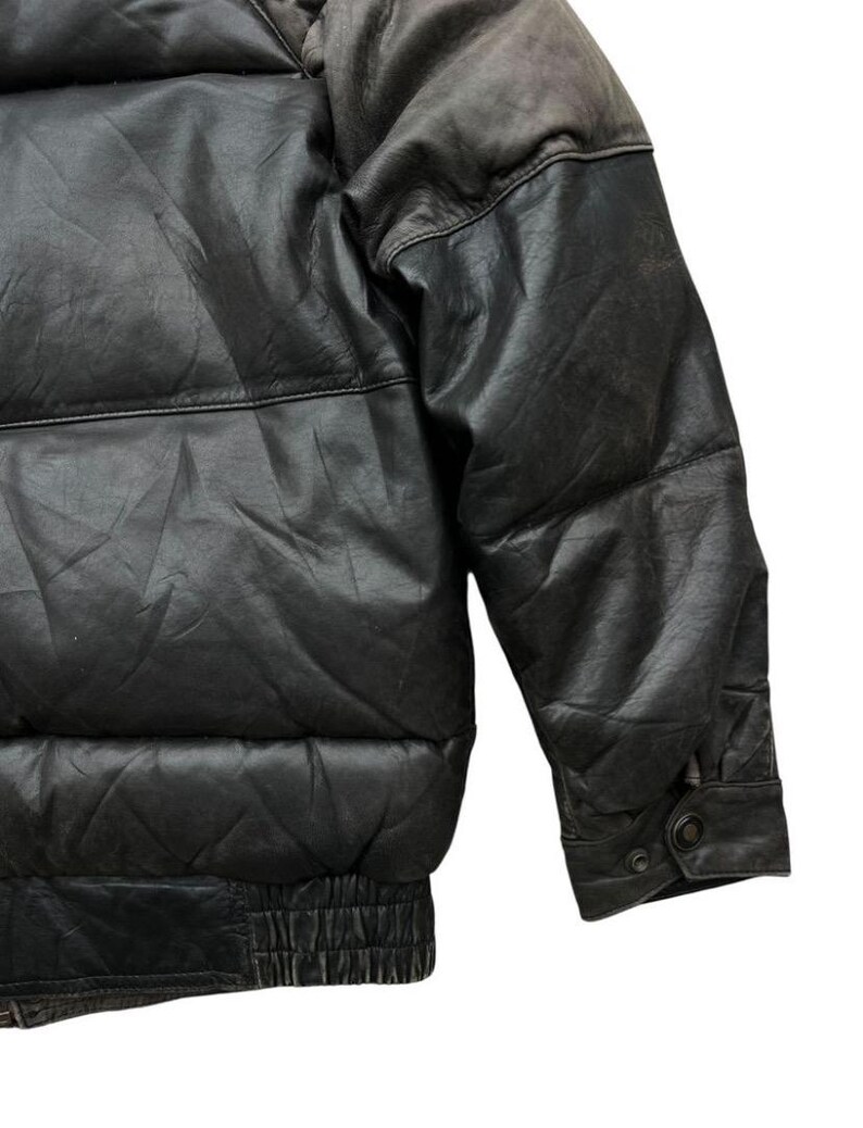 May include: Close-up of a black leather puffer jacket. The jacket features a quilted design, a ribbed hem, and a buttoned cuff. The leather has a slightly distressed appearance.