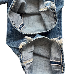 以下が含まれることがあります： 青いデニムジーンズの裾のクローズアップ。ジーンズは、ぼろぼろのほつれたエッジと、赤と白のストライプの見えるセルビッジディテールを備えています。ステッチは対照的な黄色です。