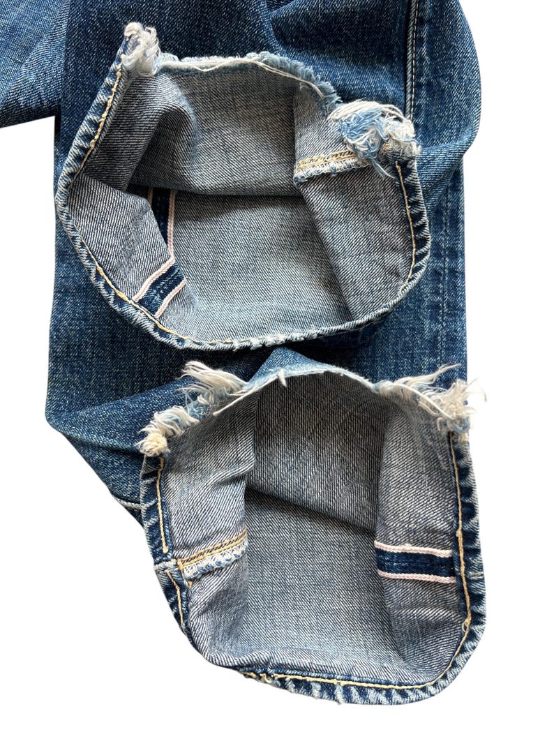 以下が含まれることがあります： 青いデニムジーンズの裾のクローズアップ。ジーンズは、ぼろぼろのほつれたエッジと、赤と白のストライプの見えるセルビッジディテールを備えています。ステッチは対照的な黄色です。