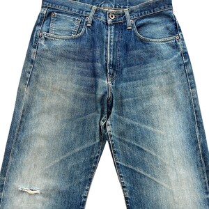 May include: Faded blue denim jeans with a distressed appearance. The jeans feature a classic five-pocket design, a button closure, and a small tear on the left leg. The denim has a worn look with lighter areas and subtle fading.