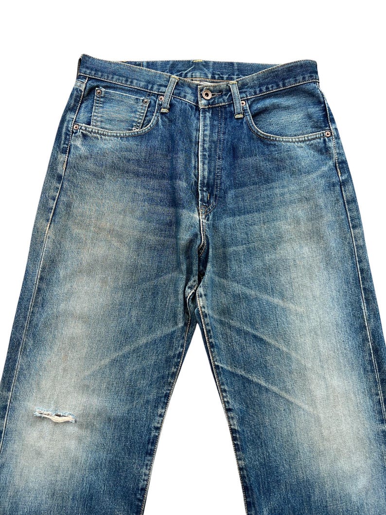 May include: Faded blue denim jeans with a distressed appearance. The jeans feature a classic five-pocket design, a button closure, and a small tear on the left leg. The denim has a worn look with lighter areas and subtle fading.