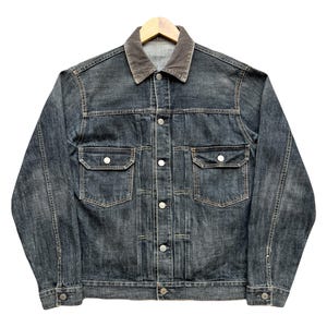 Può includere: Giacca di jeans blu scuro con colletto grigio a contrasto, chiusura con bottoni e due tasche sul petto. La giacca è appesa a una gruccia di legno su sfondo bianco. Uno stile classico.