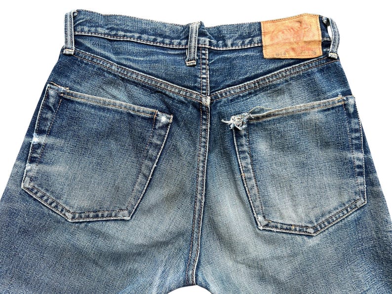 Pu&ograve; includere: Primo piano del retro di jeans blu. I jeans hanno due tasche posteriori e una toppa in pelle. Il denim &egrave; sbiadito con qualche segno di usura intorno alle tasche e ai passanti.