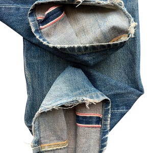 May include: Close-up of blue denim jeans with the cuffs rolled up. The inside of the cuffs reveals a red and blue stripe detail. The edges of the denim are frayed.
