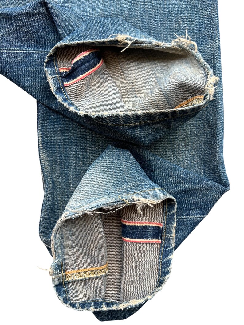 May include: Close-up of blue denim jeans with the cuffs rolled up. The inside of the cuffs reveals a red and blue stripe detail. The edges of the denim are frayed.